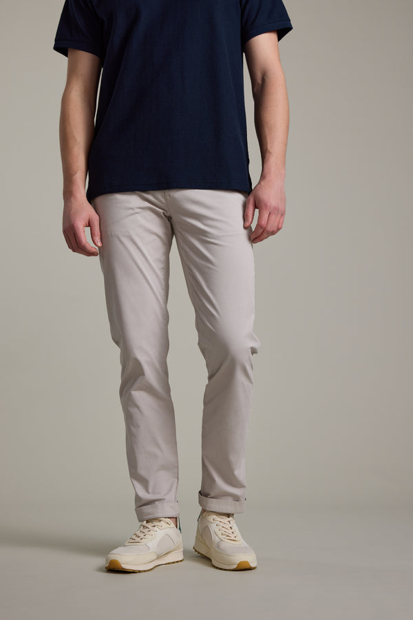 A person, head not visible, stands against a plain background wearing a short-sleeve navy t-shirt, Barkers Kingly Chino Pant in light beige, and cream sneakers.