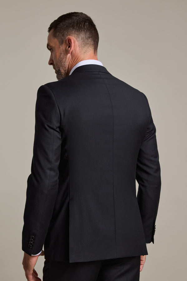 A man with short dark hair and a beard is seen from behind in a fitted black Barkers Mayfair ZQrx Suit Jacket over a white dress shirt—a sophisticated style ideal for formal events. The background is plain and light-colored.