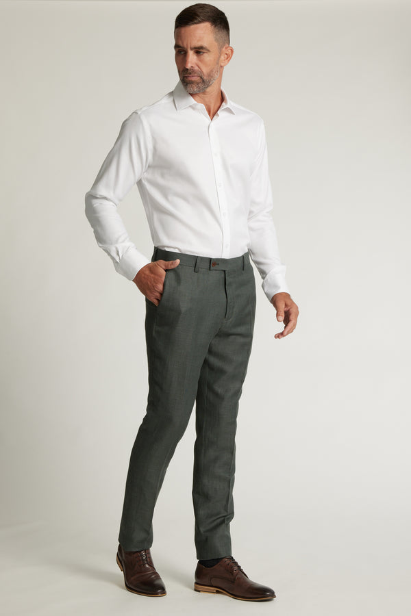 A man with short hair and a beard stands against a plain background, wearing a white shirt tucked into gray Barkers Inverness Suit Trousers with a belt and brown dress shoes, one hand in his pocket.