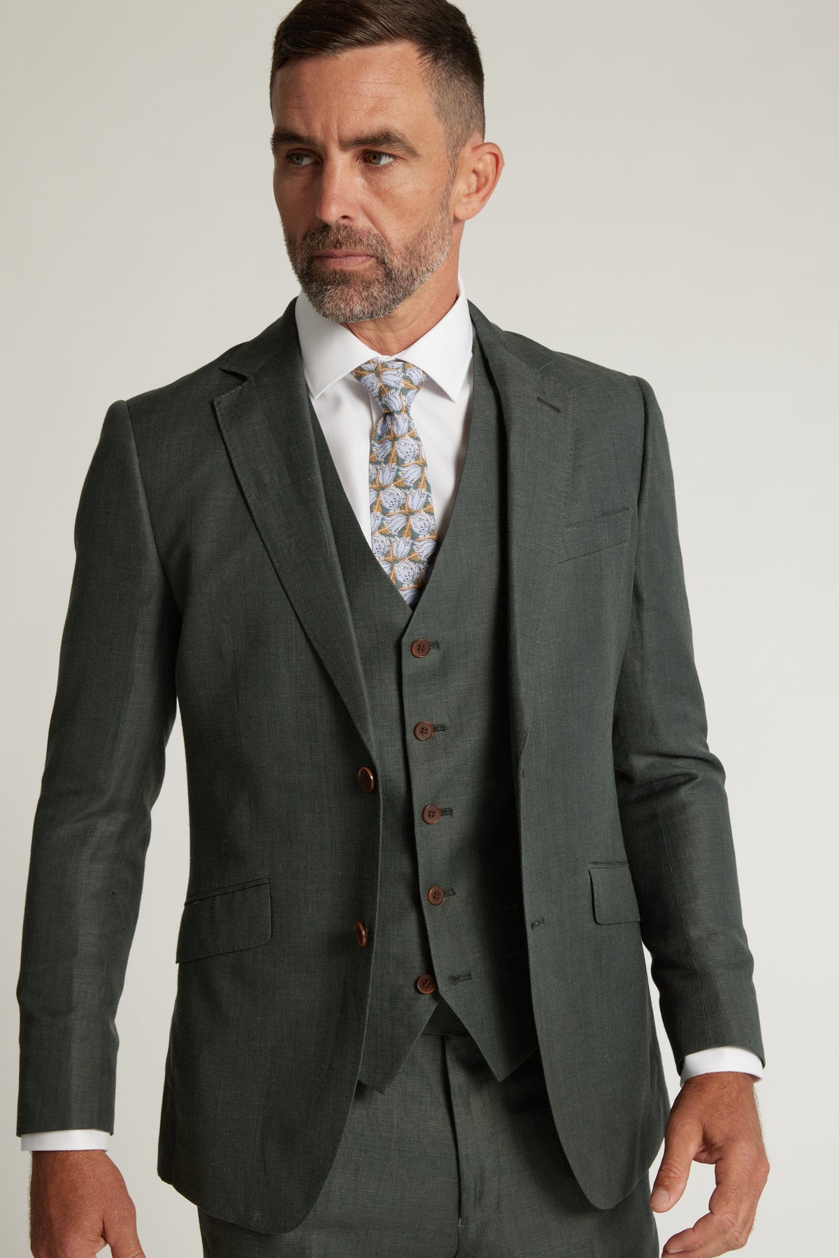 A man with short brown hair and a beard wears the Barkers Inverness Suit Jacket in sage green with a white dress shirt and patterned tie, looking to the side against a plain light background.