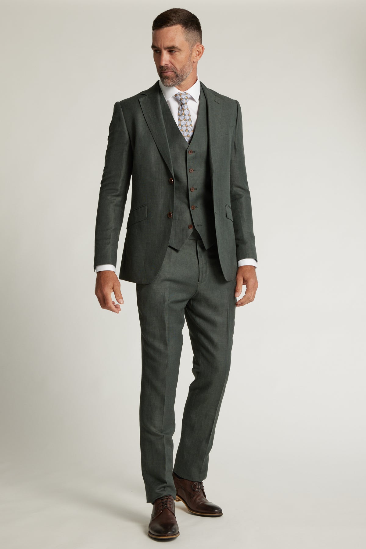 A man wears the Barkers Inverness Suit Jacket in sage green with a white shirt, patterned tie, and brown dress shoes, standing against a plain light background.