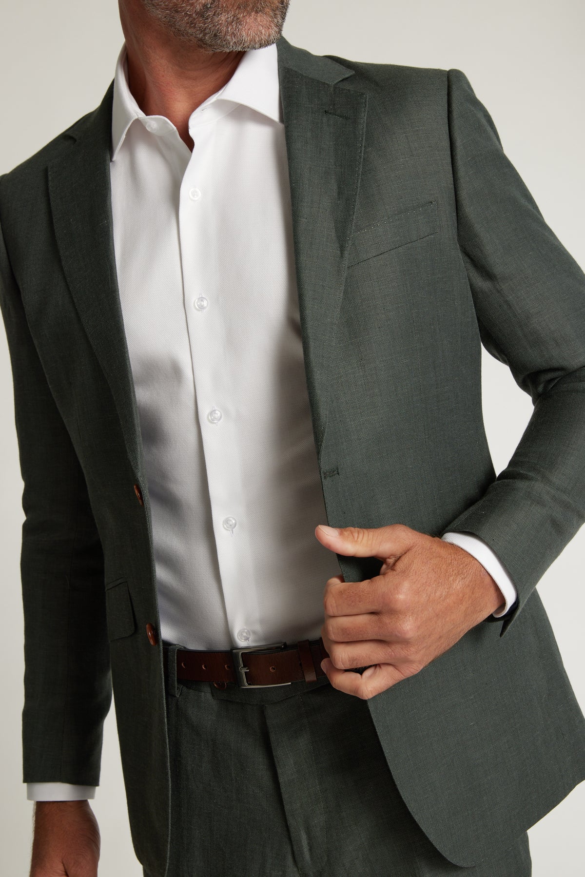 A man in a Barkers Inverness Suit Jacket in sage green over a white dress shirt adjusts his jacket with one hand, his face partly out of frame.
