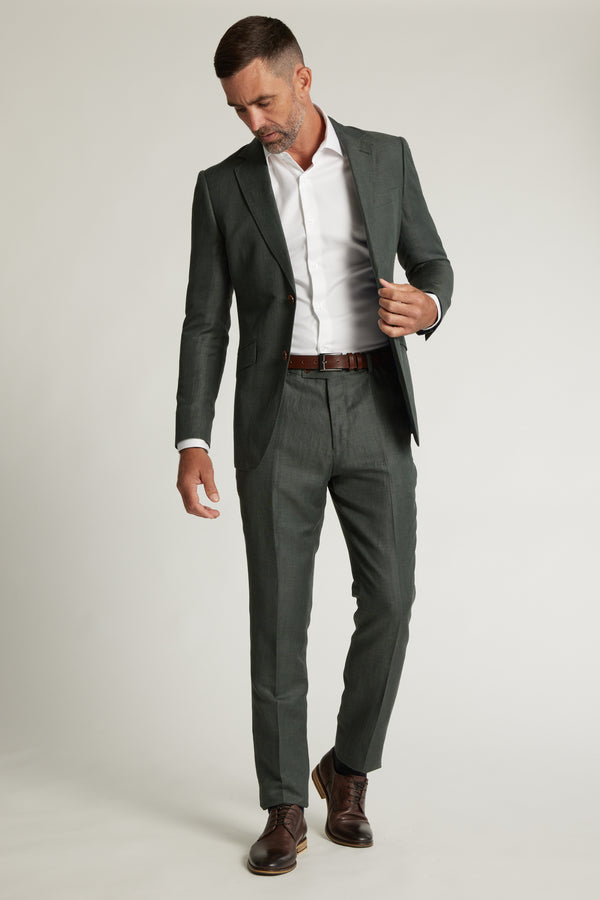 A man in Barkers' Inverness Suit Trouser, a dark green tapered fit suit, white dress shirt, and brown belt and shoes stands against a light background, looking down as he adjusts his jacket.