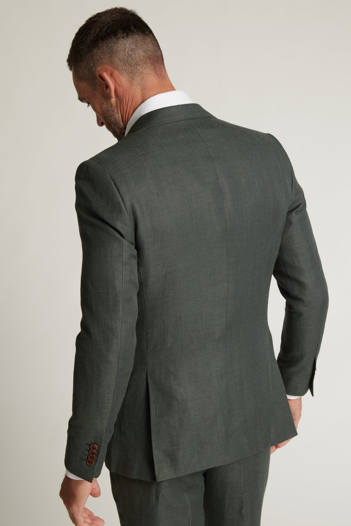 A man in a Barkers Inverness Suit Jacket in sage green stands with his back to the camera and head slightly lowered, set against a plain light background.