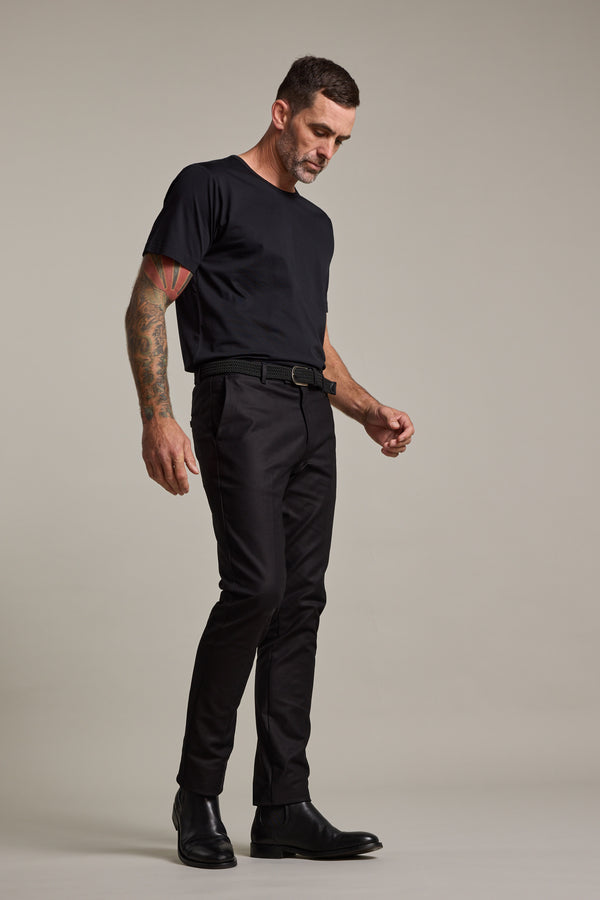 A man with short hair and arm tattoos wears a black t-shirt, Barkers Lyndon Cotton Pants, a black belt, and black boots, standing against a plain light gray background while looking down.