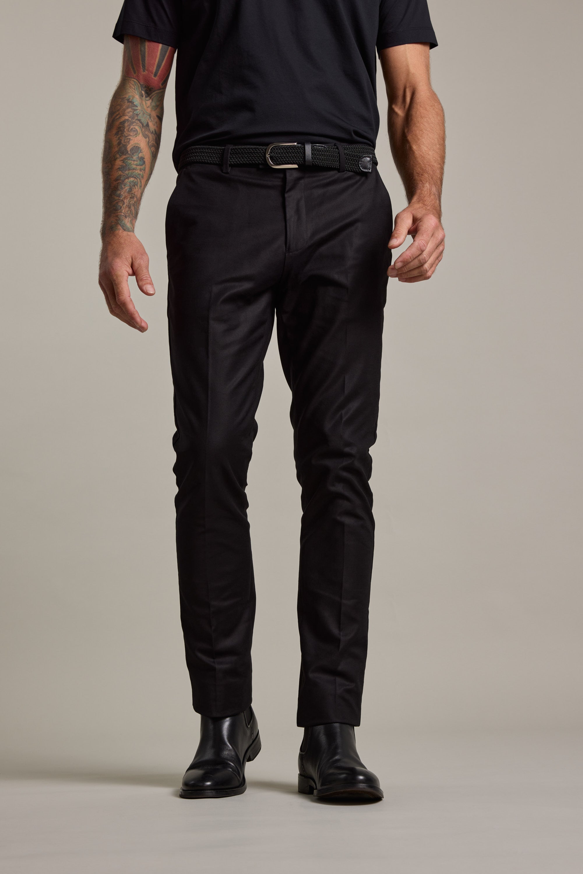 A person wears Lyndon Cotton Pants by Barkers with a black t-shirt, black belt, and black leather shoes. They stand against a plain background, their left arm showing colorful tattoos. The image is cropped at the shoulders.