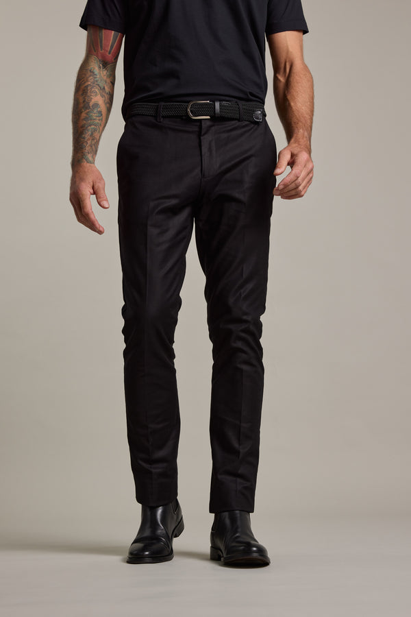 A person wears Lyndon Cotton Pants by Barkers with a black t-shirt, black belt, and black leather shoes. They stand against a plain background, their left arm showing colorful tattoos. The image is cropped at the shoulders.