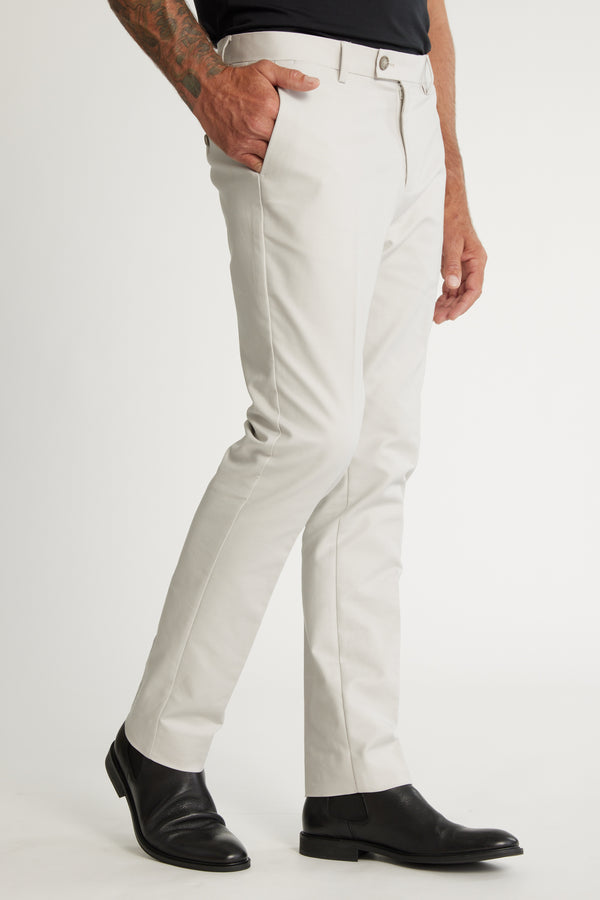 A person wearing Barkers Lyndon Cotton Pants in a slim fit with a belt, paired with black dress shoes and a black shirt, stands with one hand in their pocket—showcasing a refined smart casual look. Only the lower half is visible.