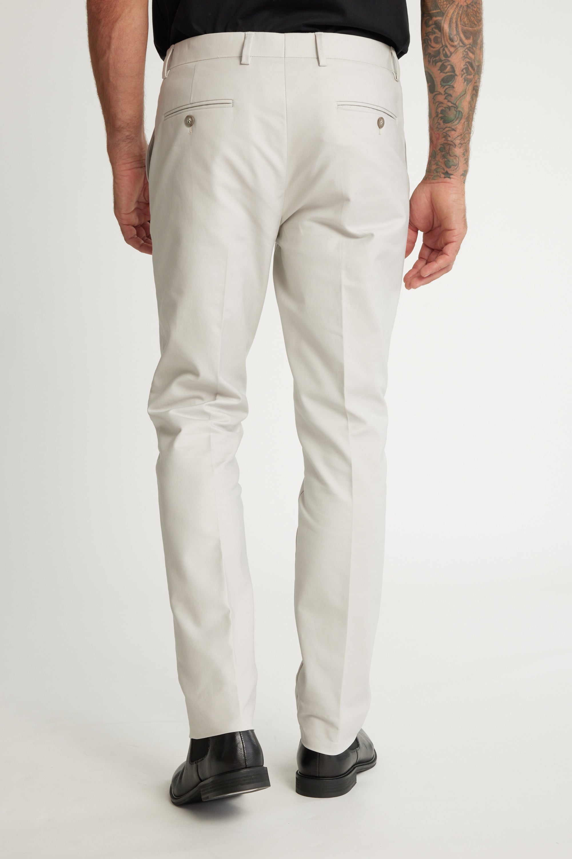 A man faces away in a smart casual look, wearing Barkers Lyndon Cotton Pants in a light color with a black belt, black shoes, and black shirt. His tattooed right arm is visible against a plain white background.