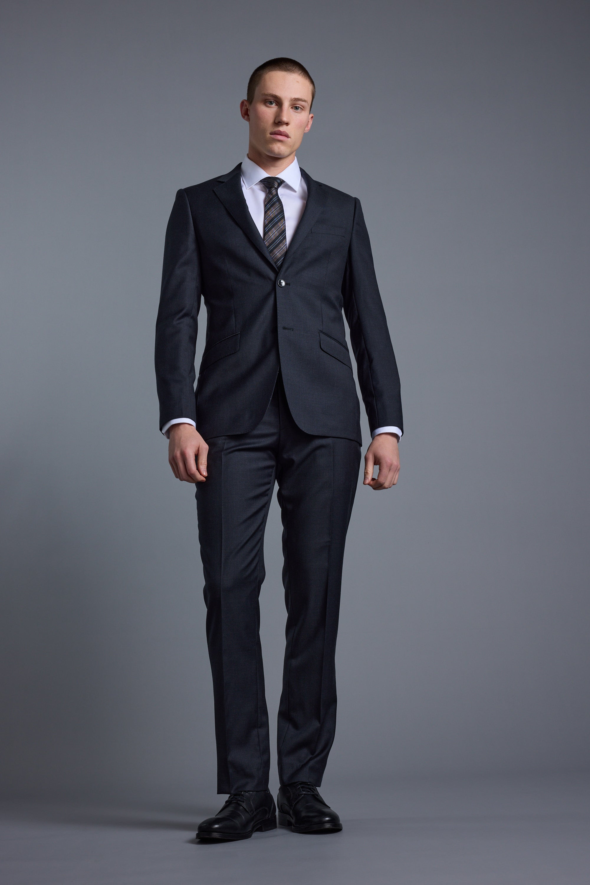 A man stands against a gray background, wearing the Barkers Mayfair ZQ Suit Jacket made from Merino Wool, paired with a white shirt, striped tie, and black dress shoes. He faces forward with a neutral expression and relaxed arms at his sides.
