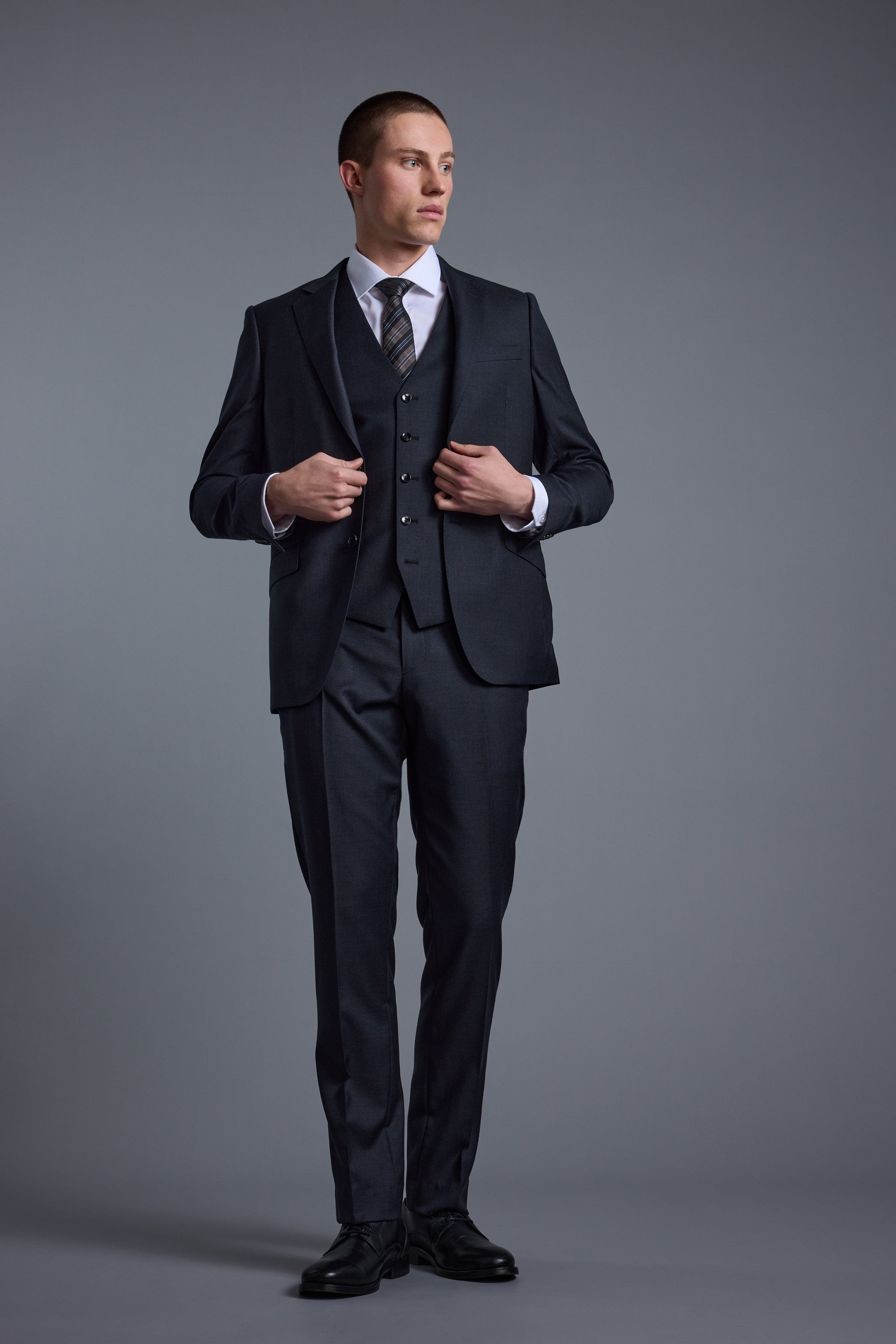 A man wearing the Barkers Mayfair ZQ Suit Jacket, crafted from New Zealand Merino, stands against a plain gray background, looking to his left as he adjusts his jacket with both hands.