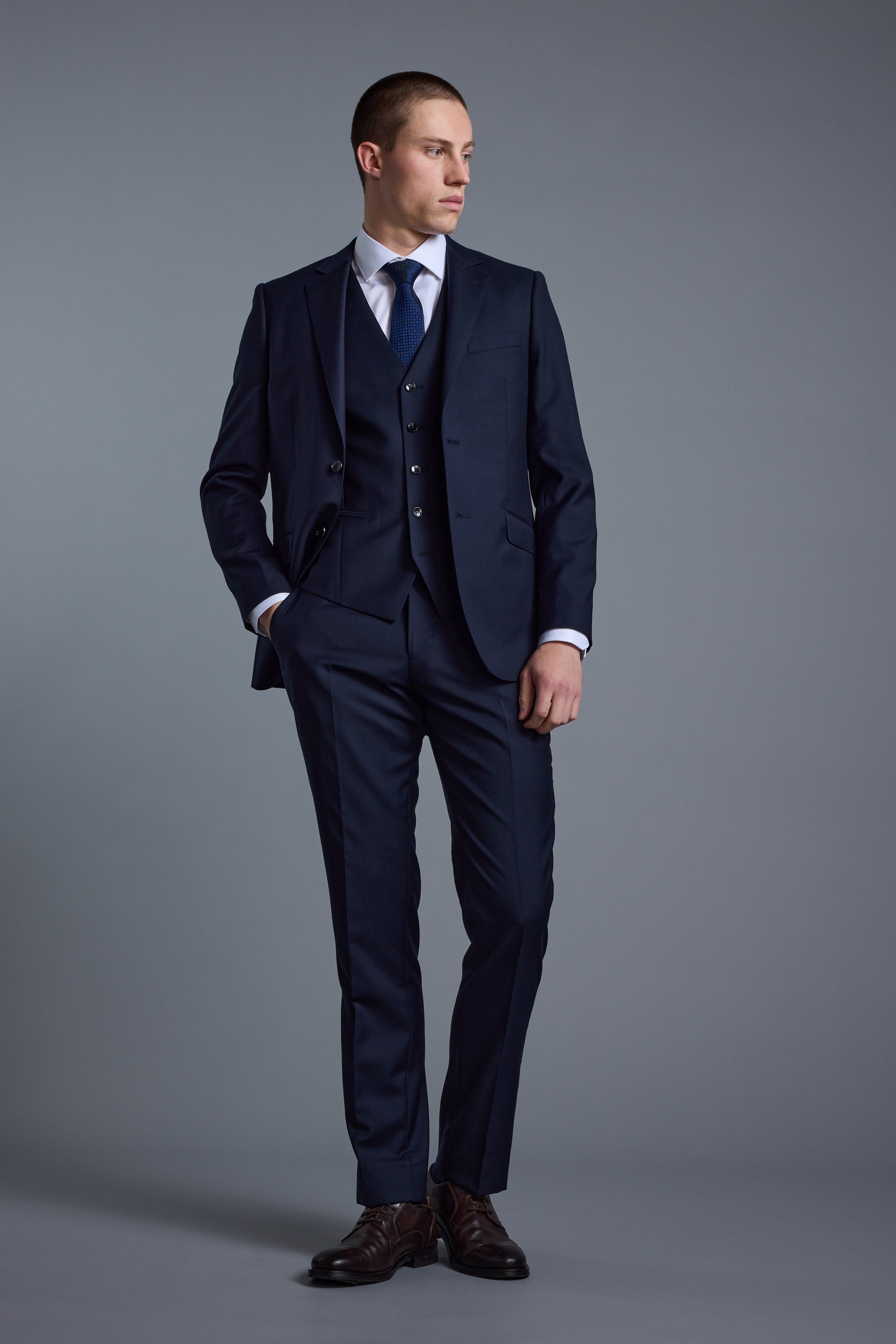Man in Barkers Mayfair ZQrx Suit Trouser, white shirt, and blue tie stands before a gray background.