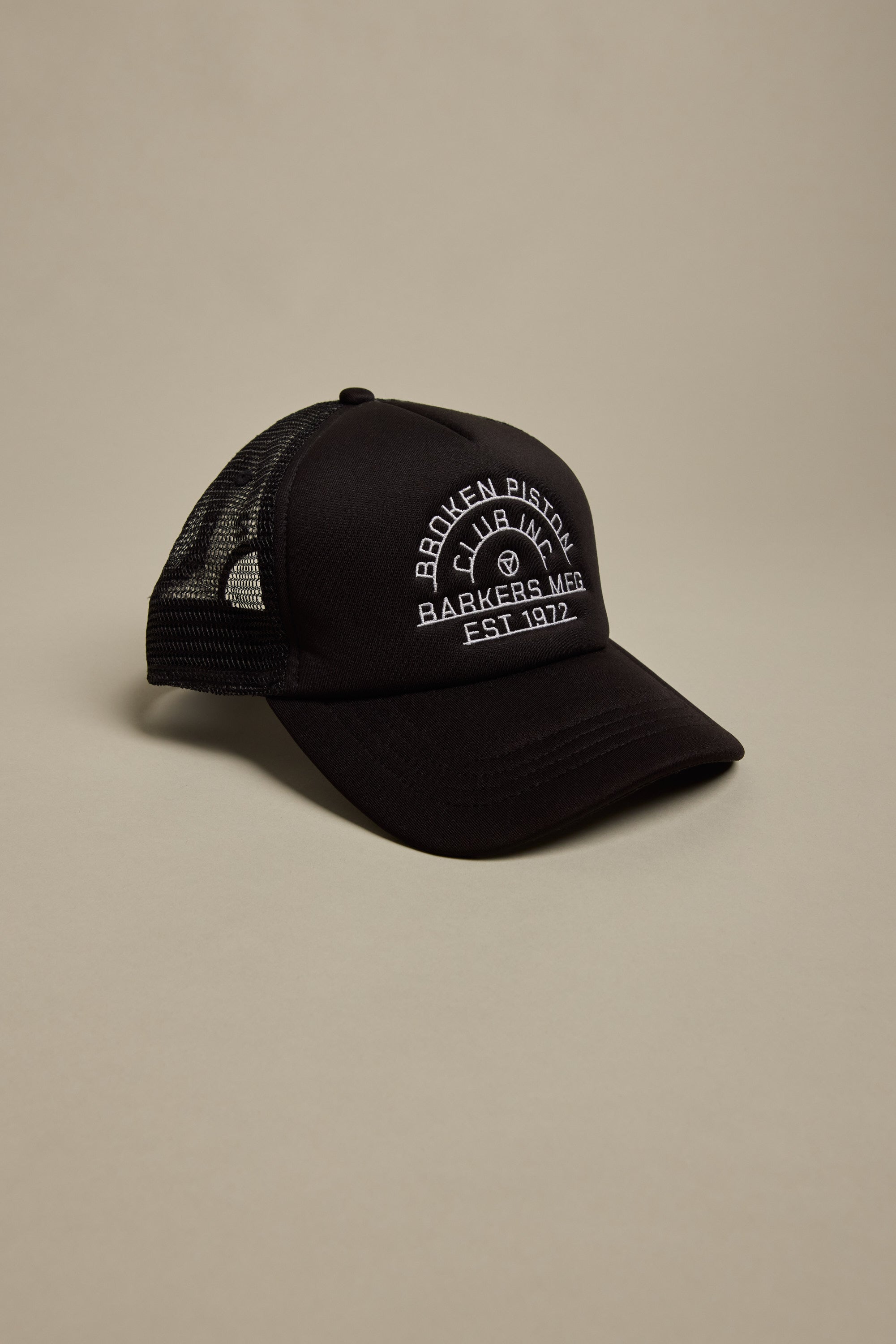The Barkers Piston Club Trucker Cap is a black mesh cap with a curved brim, adjustable fit, and a white embroidered "Broken Piston Barkers M&R Est. 1972" logo with piston graphic on the front, shown against a neutral background.