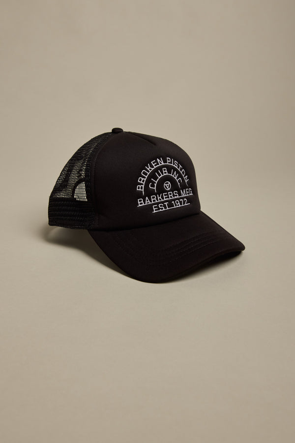 The Barkers Piston Club Trucker Cap is a black mesh cap with a curved brim, adjustable fit, and a white embroidered "Broken Piston Barkers M&R Est. 1972" logo with piston graphic on the front, shown against a neutral background.