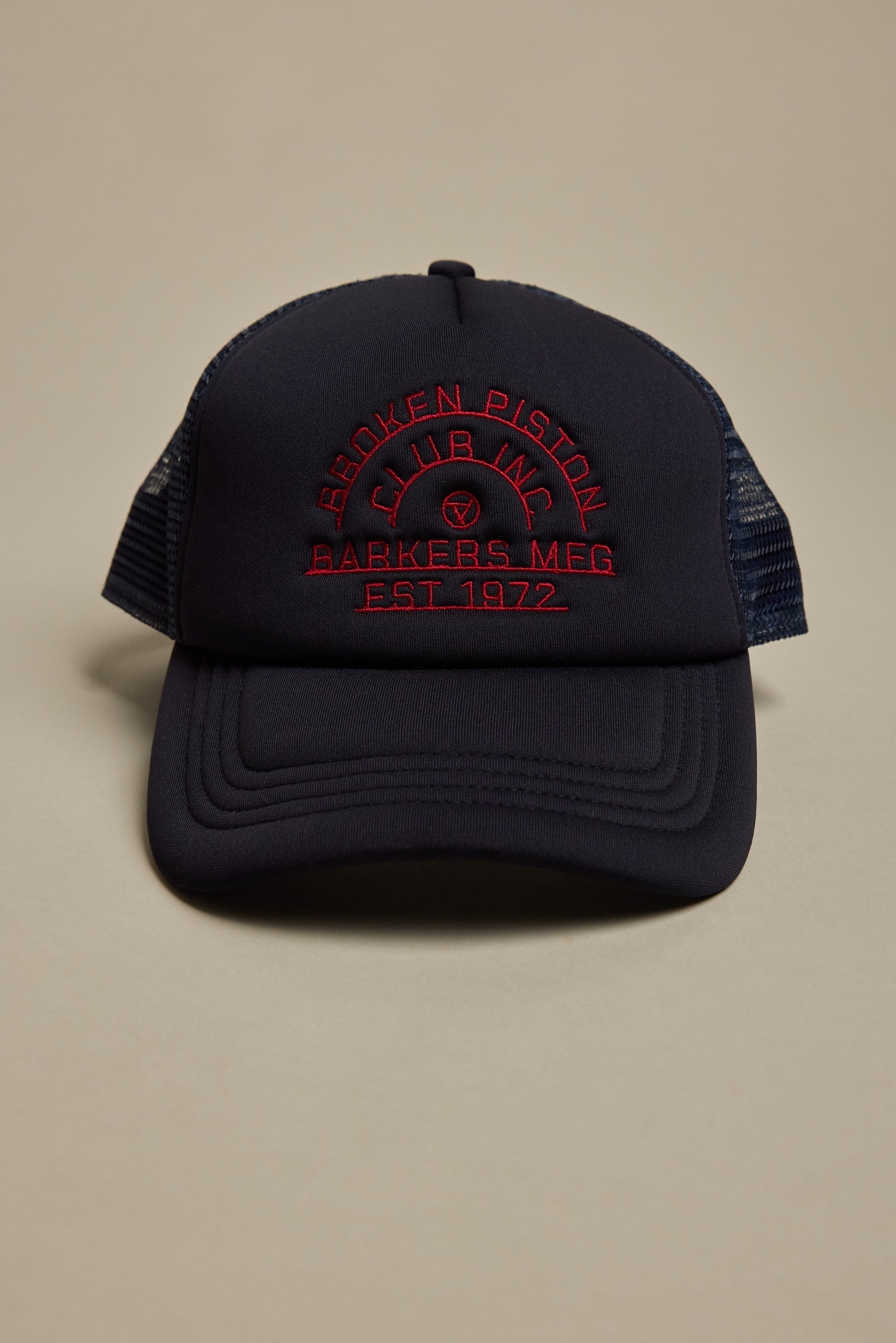 The Piston Club Trucker Cap by Barkers is a black mesh hat with an adjustable fit and a red embroidered BROKEN PISTON CLUB INC. BAKERS MEG EST. 1972 logo on the front, shown against a neutral beige background.