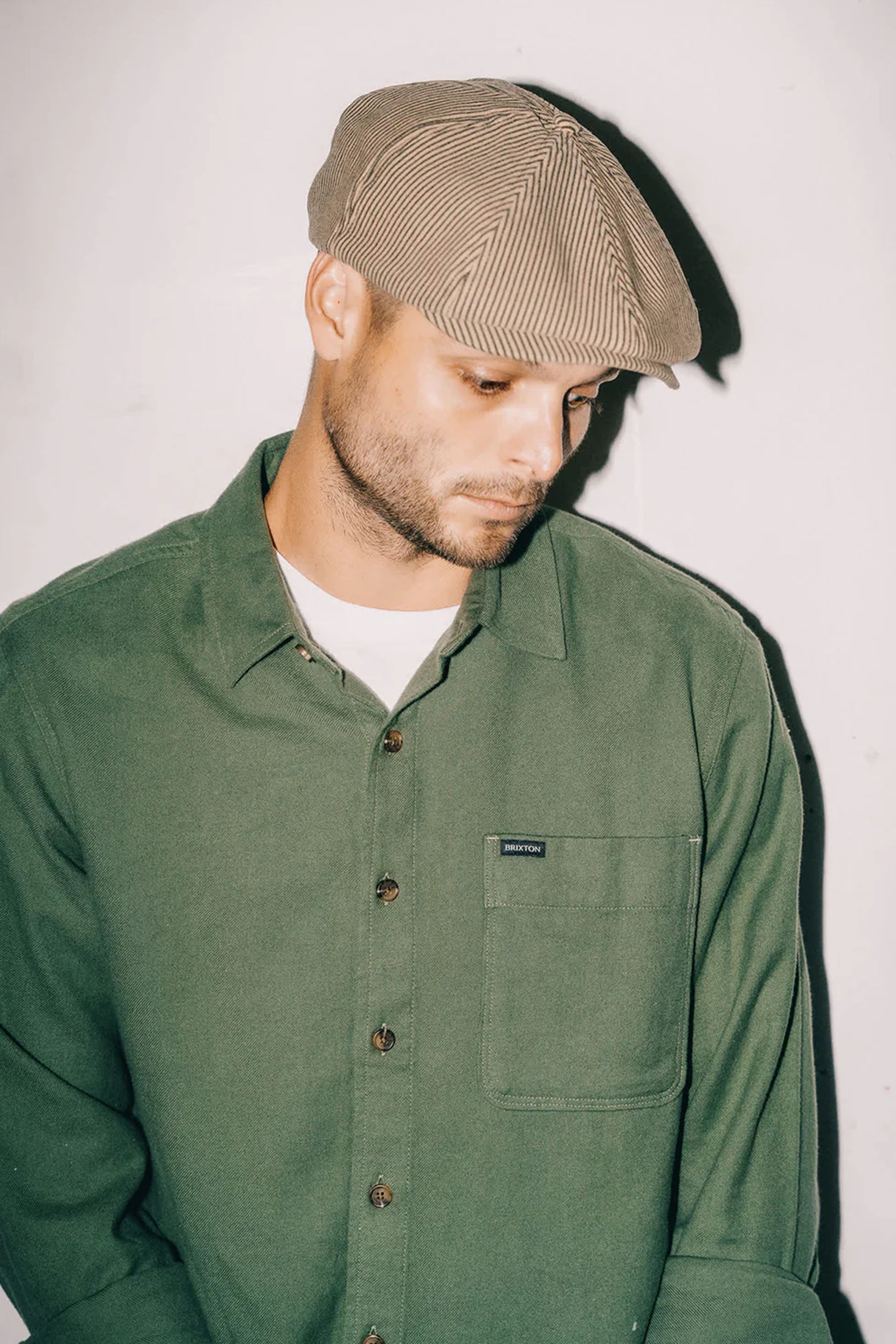 A man in a green button-up shirt and a beige Brixton Brood Snap cap by Brixton looks down with a neutral expression against a plain light-colored background.