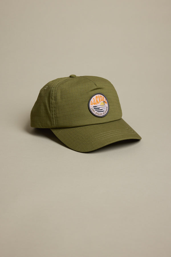 The Barkers Longboard Cap by Barkers is an olive green snapback featuring a round, colorful embroidered "CACKLE" patch with a sunset over mountains on the front. It is displayed on a light beige background.
