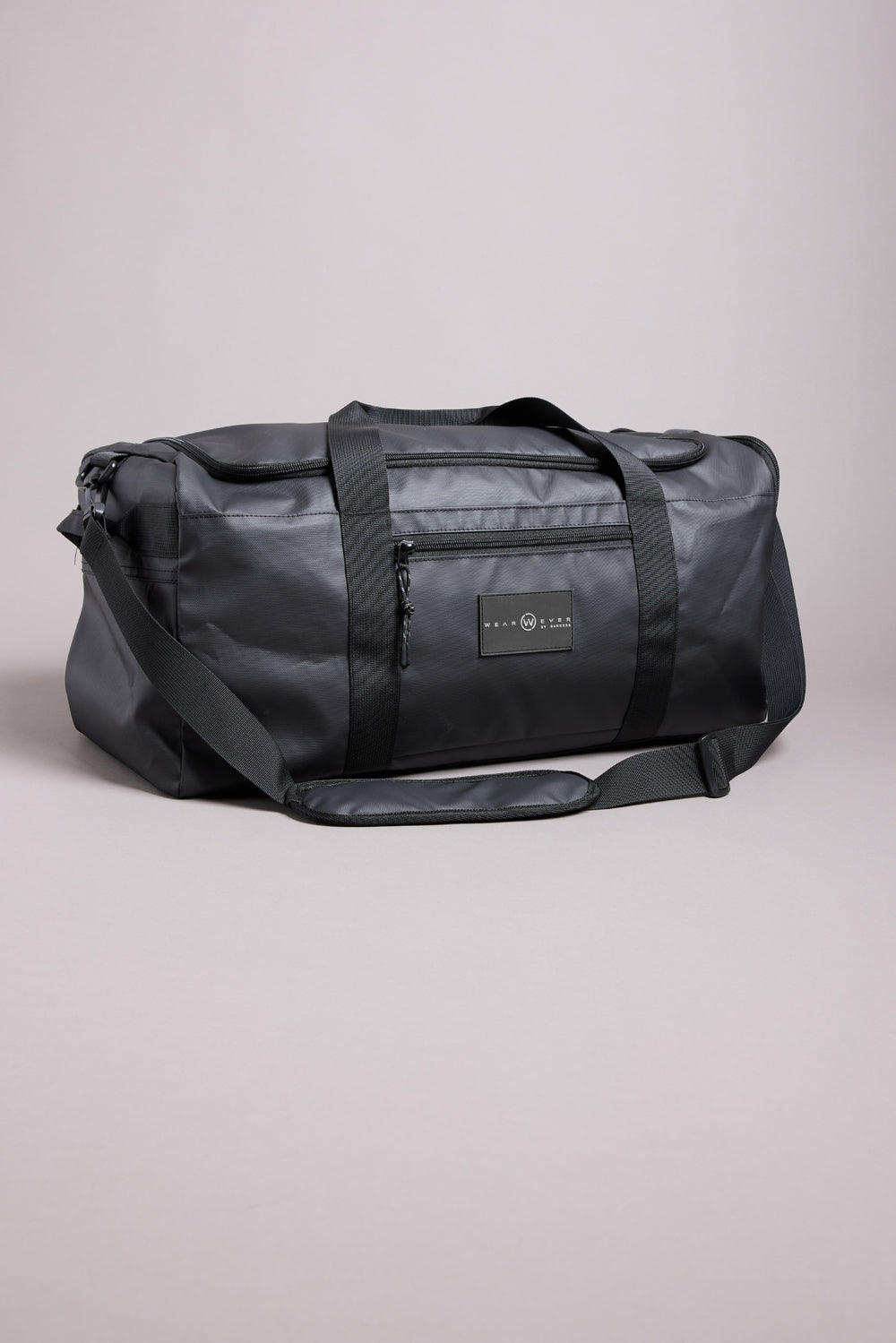 The WearEver Transit Holdall by WearEver is a black, water-resistant duffel bag featuring two handles, a shoulder strap, sneaker pocket, and zippered front pocket, shown against a neutral light gray background.