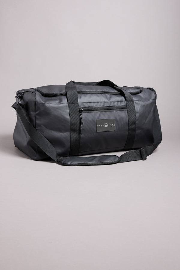 The WearEver Transit Holdall by WearEver is a black, water-resistant duffel bag featuring two handles, a shoulder strap, sneaker pocket, and zippered front pocket, shown against a neutral light gray background.