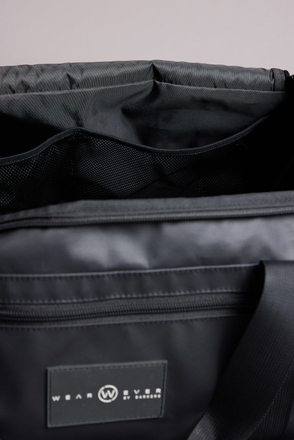 Close-up of the WearEver Transit Holdall’s open interior shows mesh compartments and a sneaker pocket. The black, water-resistant exterior has a zip pocket and displays the WearEver logo on a circular label.