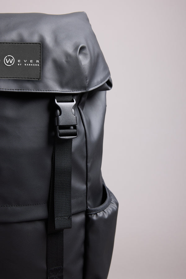 Close-up of the sleek black WearEver Transit Backpack, featuring a flap cover, water-resistant zips, black buckle strap, and a small "WearEver" logo patch on the upper left side against a plain light gray background.