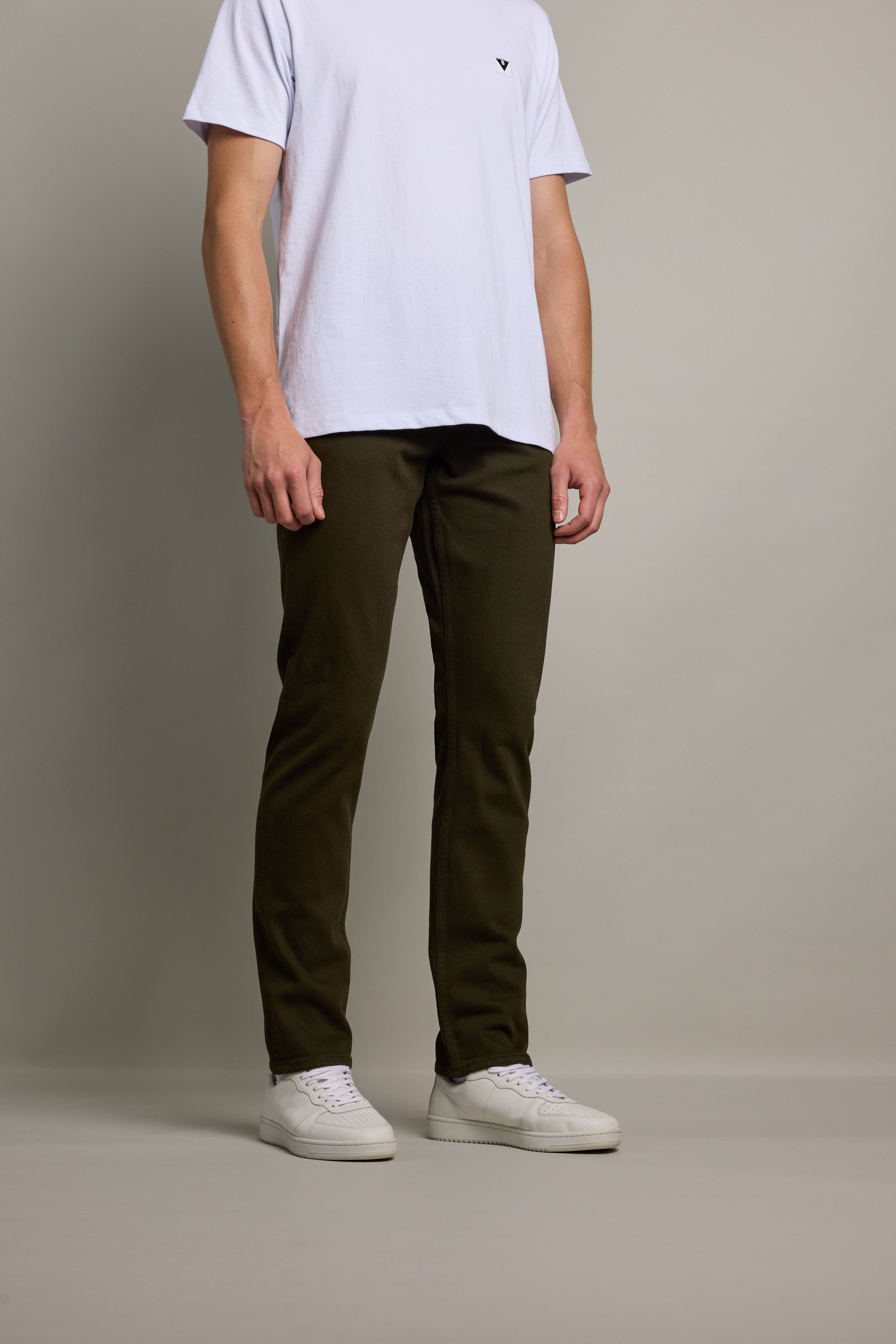 A person wearing a white T-shirt and Barkers Rambler Slim Coloured Jean in dark olive green regenerative cotton stands against a plain light gray background with their face not visible.