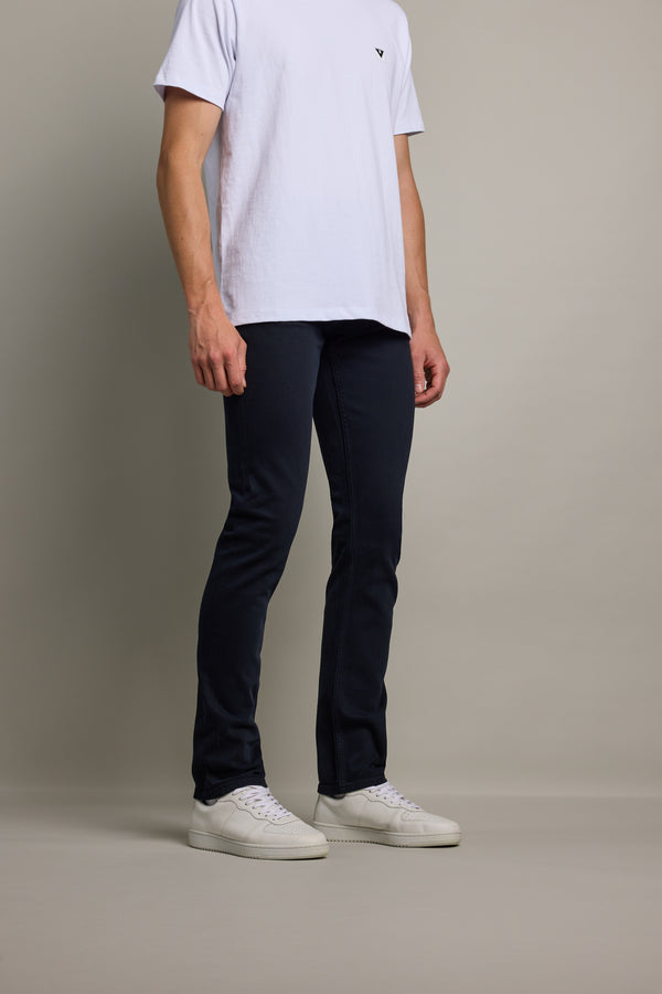A person wearing a white t-shirt, Barkers Rambler Slim Coloured Jean made from regenerative cotton by Saitex Vietnam, and white sneakers stands against a plain light gray background. The person’s face is not visible.
