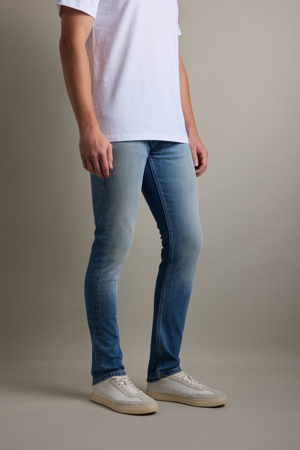 Person wearing Barkers Rambler Denim Slim Jean, white t-shirt, and white sneakers indoors.