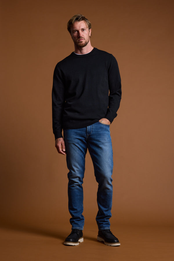 A man with short light hair wears a black Barkers Skyland ZQrx Merino Crew, made from sustainable New Zealand merino wool, paired with blue jeans and black shoes, standing against a plain brown background with one hand in his pocket.