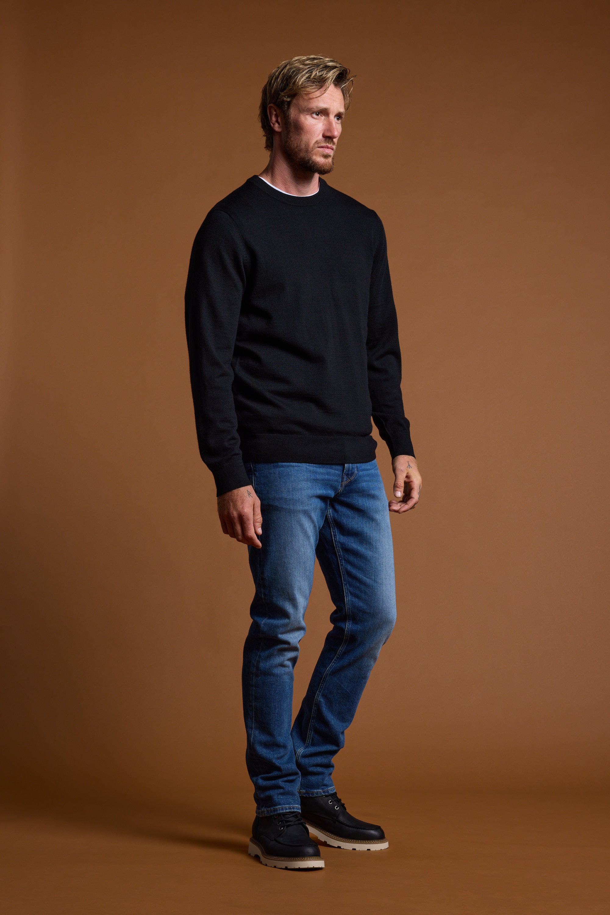 A man with short blond hair and a beard stands against a brown background, wearing the Barkers Skyland ZQrx Merino Crew sweater, blue jeans, and black shoes. He looks to the side with a neutral expression, showcasing the sustainable merino knit.