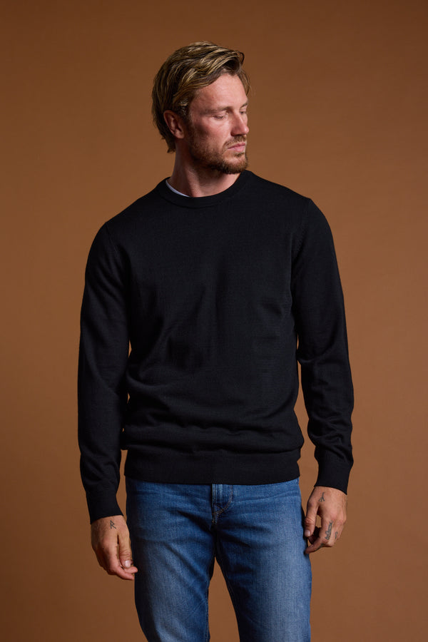 A blond man with a beard wears the Barkers Skyland ZQrx Merino Crew, a sustainable merino wool sweater, with blue jeans. He stands against a brown background, looking left with a neutral expression.