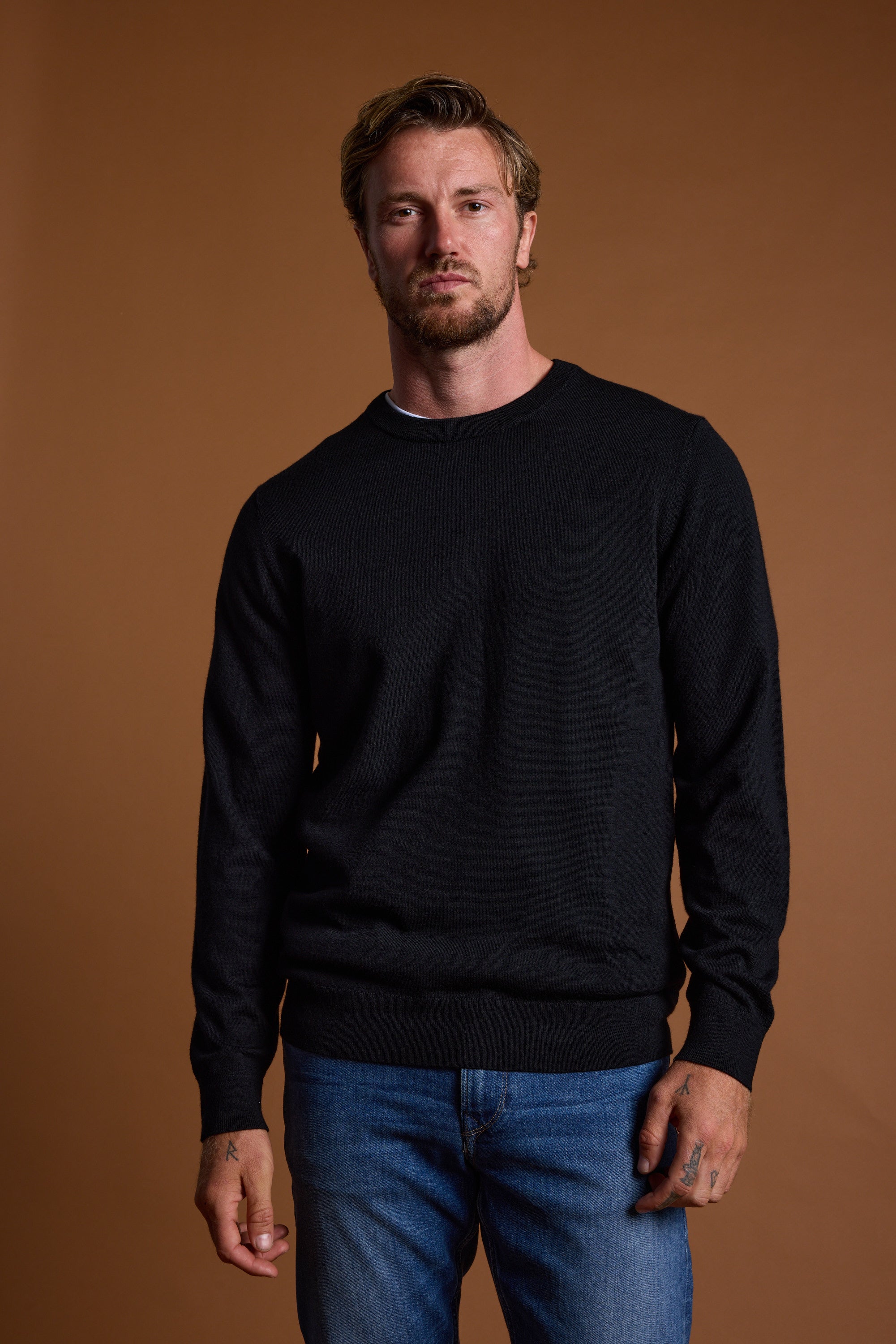 A man with short, light brown hair and a beard stands against a brown background, wearing the Barkers Skyland ZQrx Merino Crew sweater and blue jeans. He has a neutral expression and his left hand is slightly clenched.