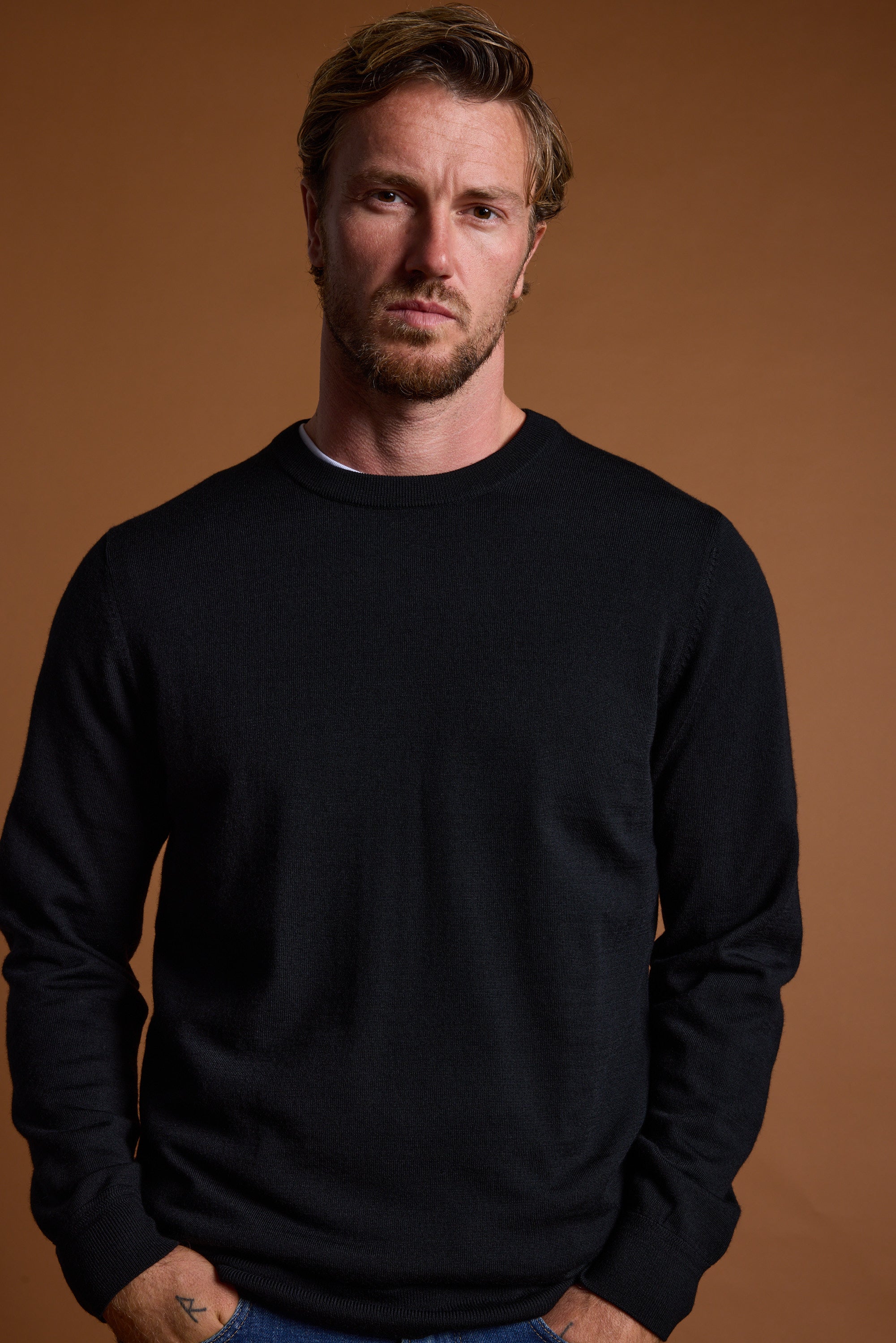A man with light brown hair and a beard, wearing a black Barkers Skyland ZQrx Merino Crew sweater made from sustainable merino wool, stands against a plain brown background with a neutral expression and his hands in his pockets.