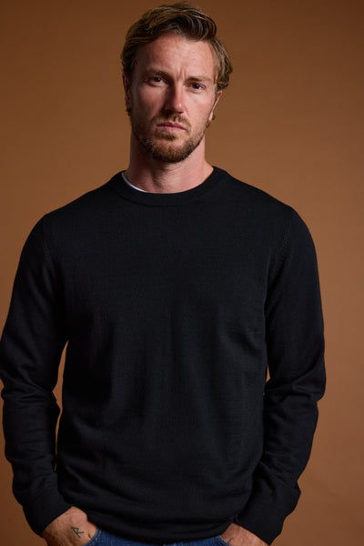 A man with short light brown hair and a beard wears the Barkers Skyland ZQrx Merino Crew, made from ethically sourced wool, standing against a plain brown background with his hands in his pockets, looking at the camera.