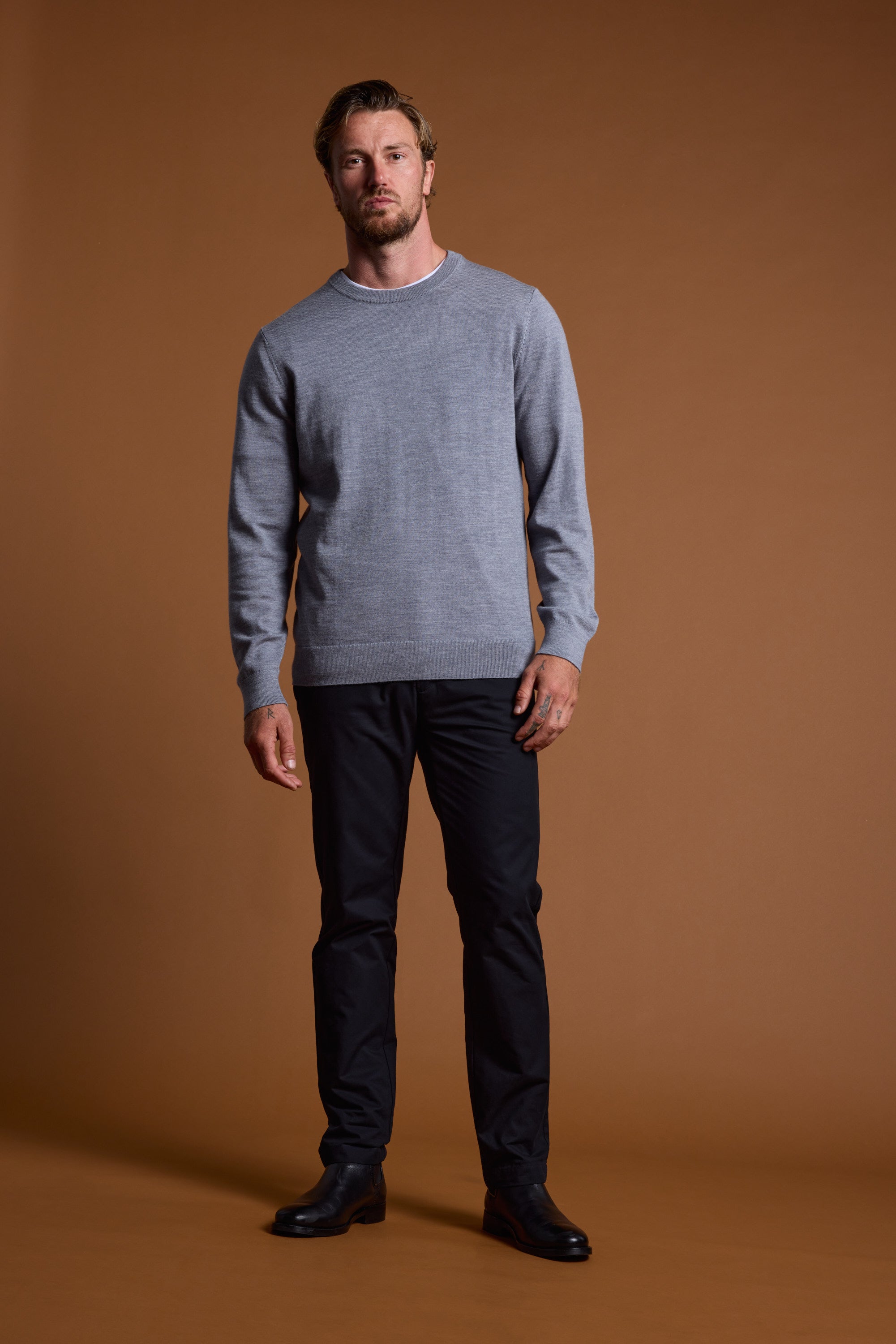 A man with short hair and a beard stands against a brown background, wearing the Barkers Skyland ZQrx Merino Crew made from sustainable New Zealand merino wool, along with black pants and black shoes. He faces forward, arms relaxed at his sides.