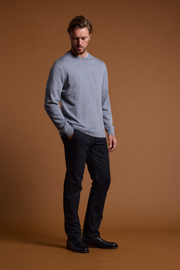 A man stands against a plain brown backdrop, wearing the Barkers Skyland ZQrx Merino Crew in light gray, with black pants and shoes. One hand is in his pocket as he looks down thoughtfully.