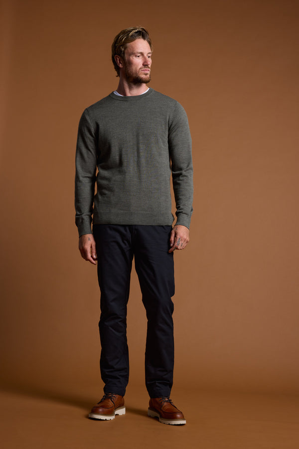 A man with short hair and a beard stands against a brown background, wearing the Barkers Skyland ZQrx Merino Crew made from ethically sourced New Zealand merino wool. He looks left with a neutral expression, paired with dark pants and brown shoes.