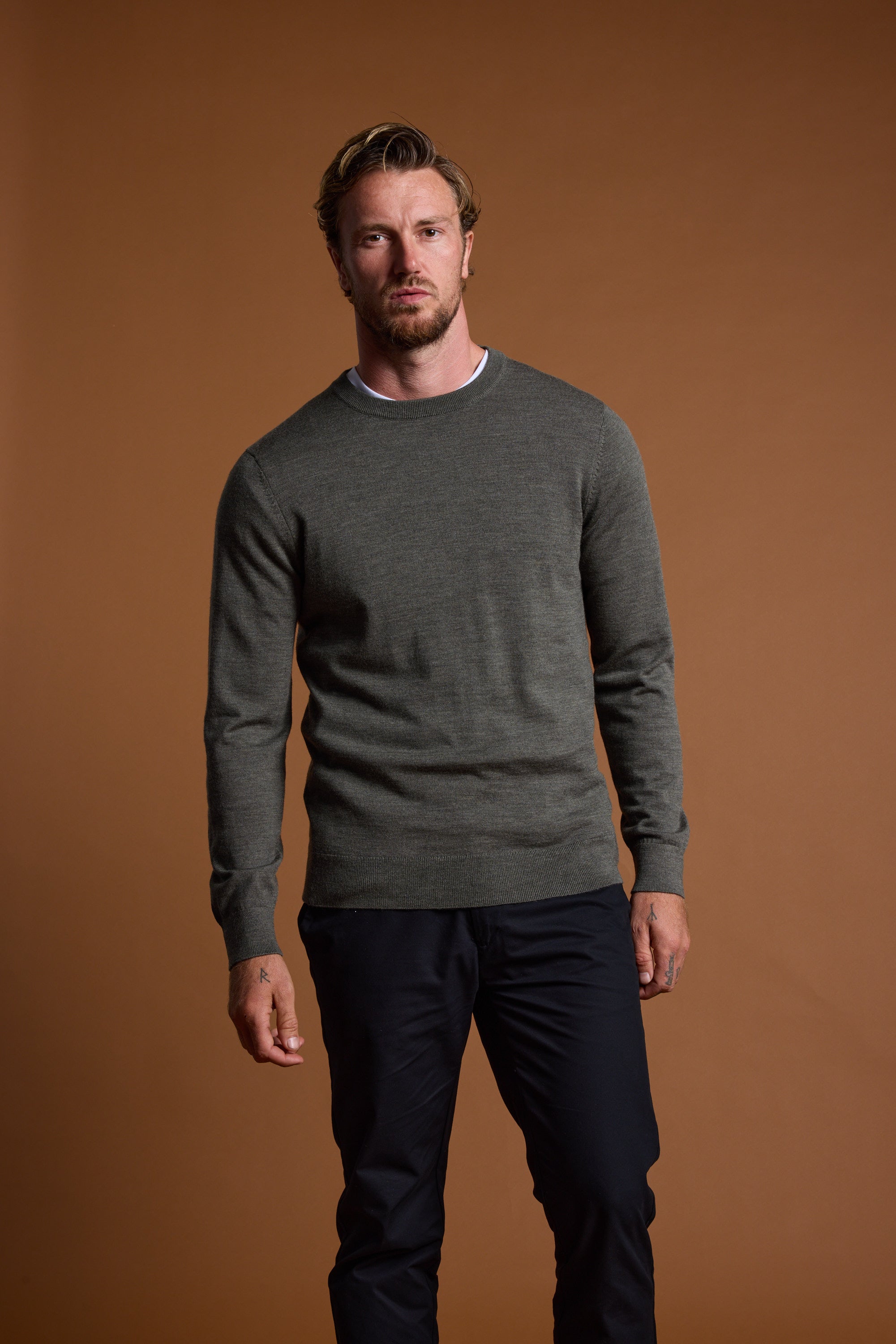 A man with short, wavy hair and a trimmed beard stands against a plain brown background, wearing the Barkers Skyland ZQrx Merino Crew sweater over a white T-shirt and dark pants.