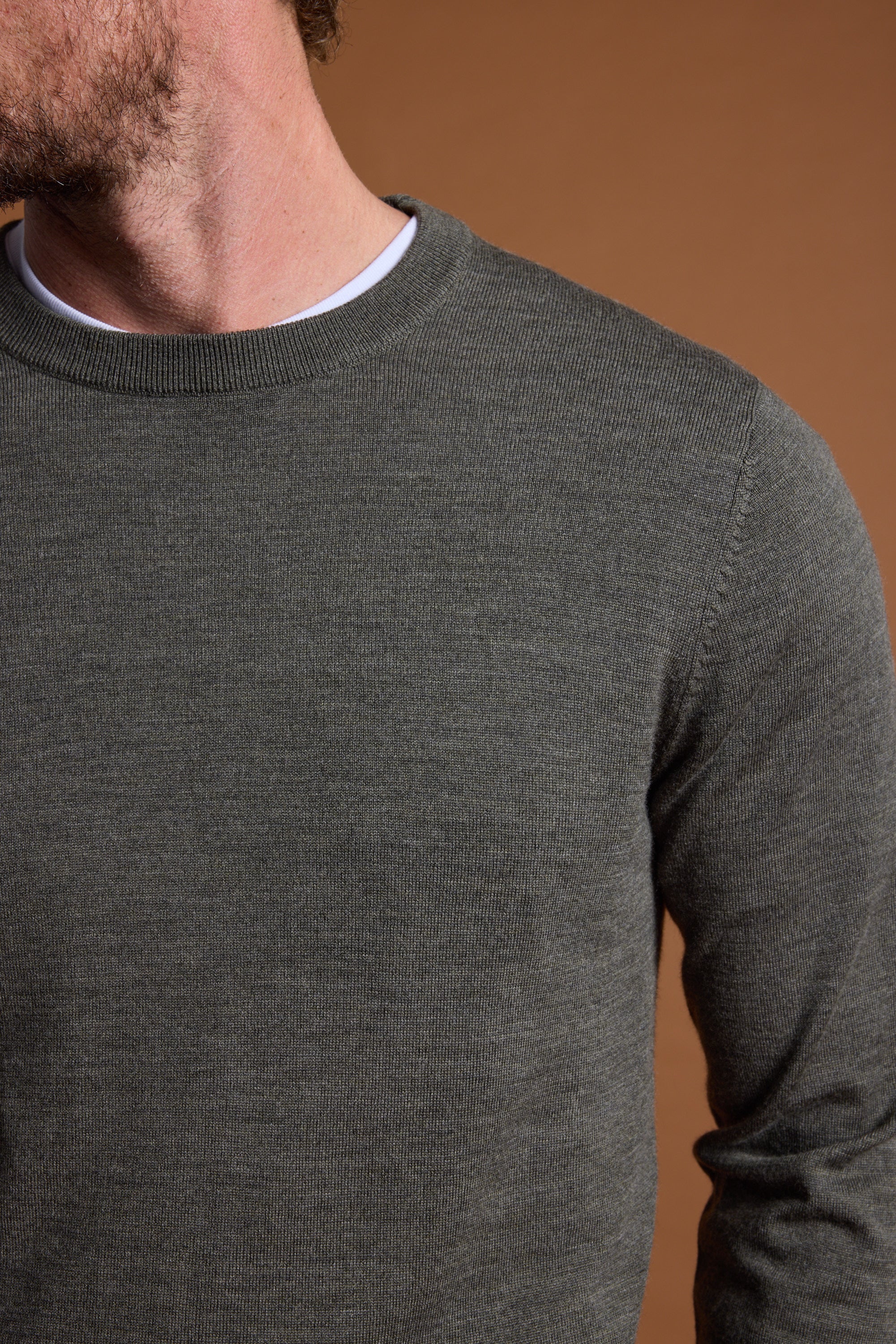 A close-up shows a person wearing the Barkers Skyland ZQrx Merino Crew in dark grey over a white t-shirt, set against a brown background. Only their lower face and neck are visible, with the head partially out of frame.