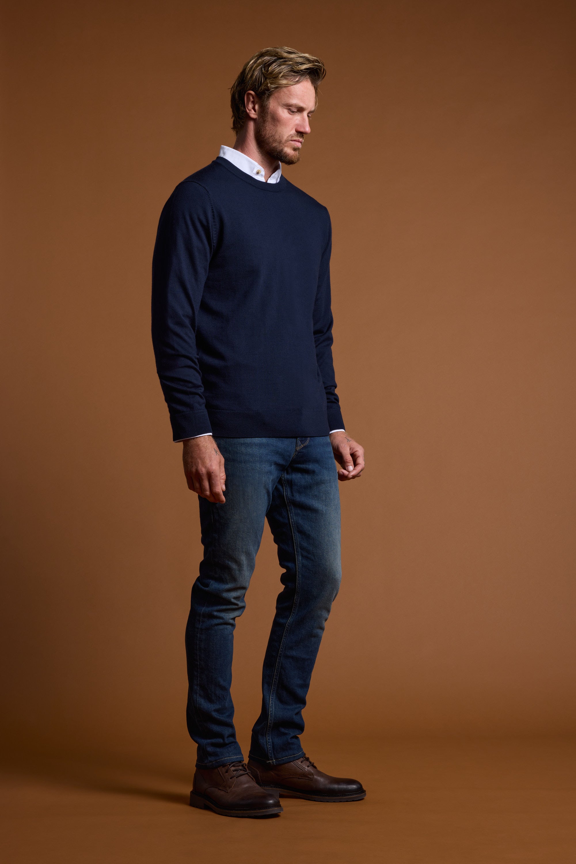 A man with light brown hair and a beard stands against a brown background, wearing the Barkers Skyland ZQrx Merino Crew over a white shirt, blue jeans, and brown shoes, looking downward with a neutral expression.