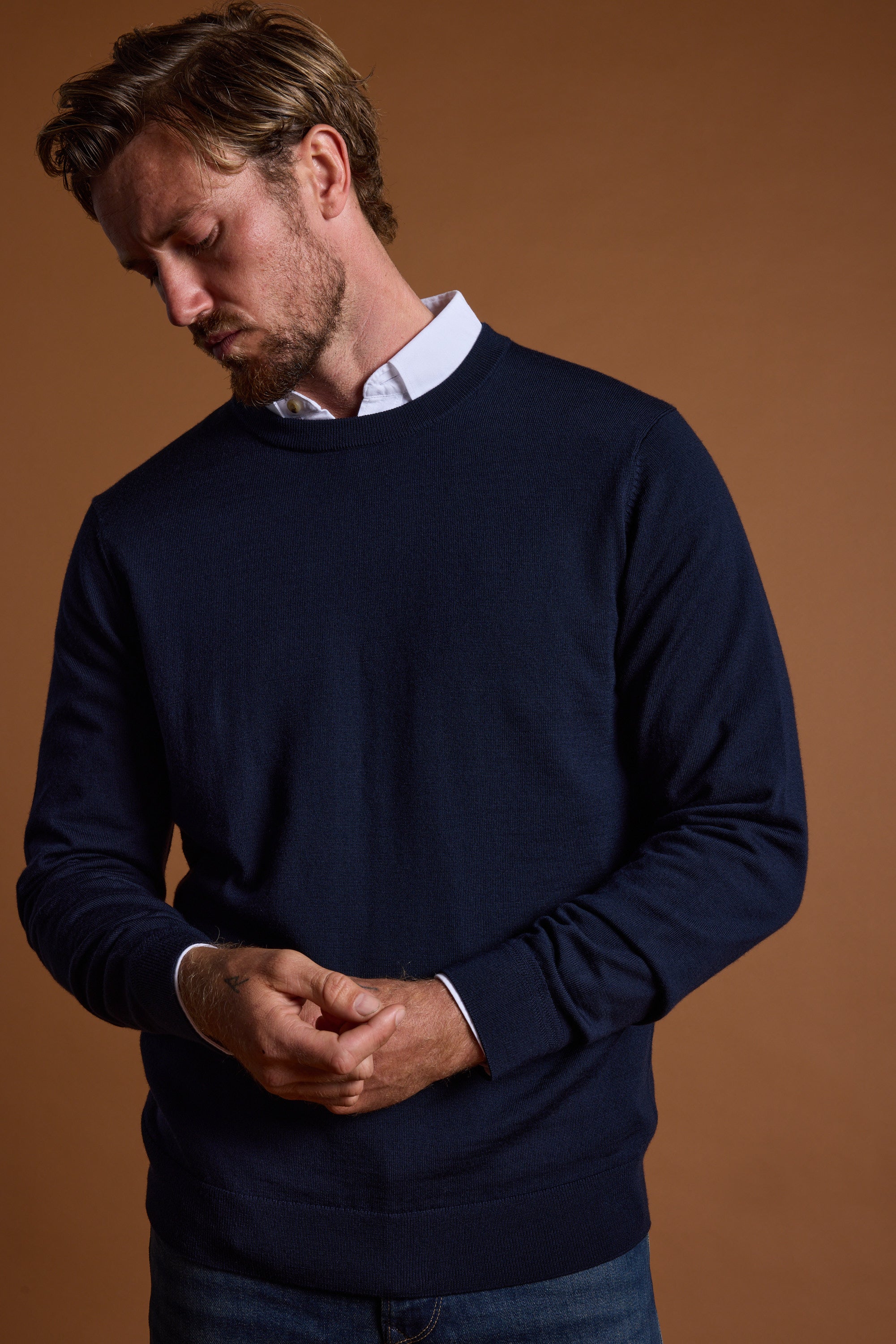 A man with light brown hair and a beard wears the Barkers Skyland ZQrx Merino Crew sweater, navy blue over a white collared shirt. He looks down with hands loosely clasped, standing against a plain brown background.