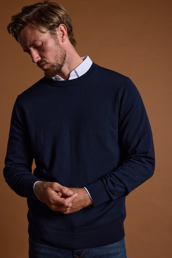 A man with light brown hair and a beard wears the Barkers Skyland ZQrx Merino Crew sweater, navy blue over a white collared shirt. He looks down with hands loosely clasped, standing against a plain brown background.