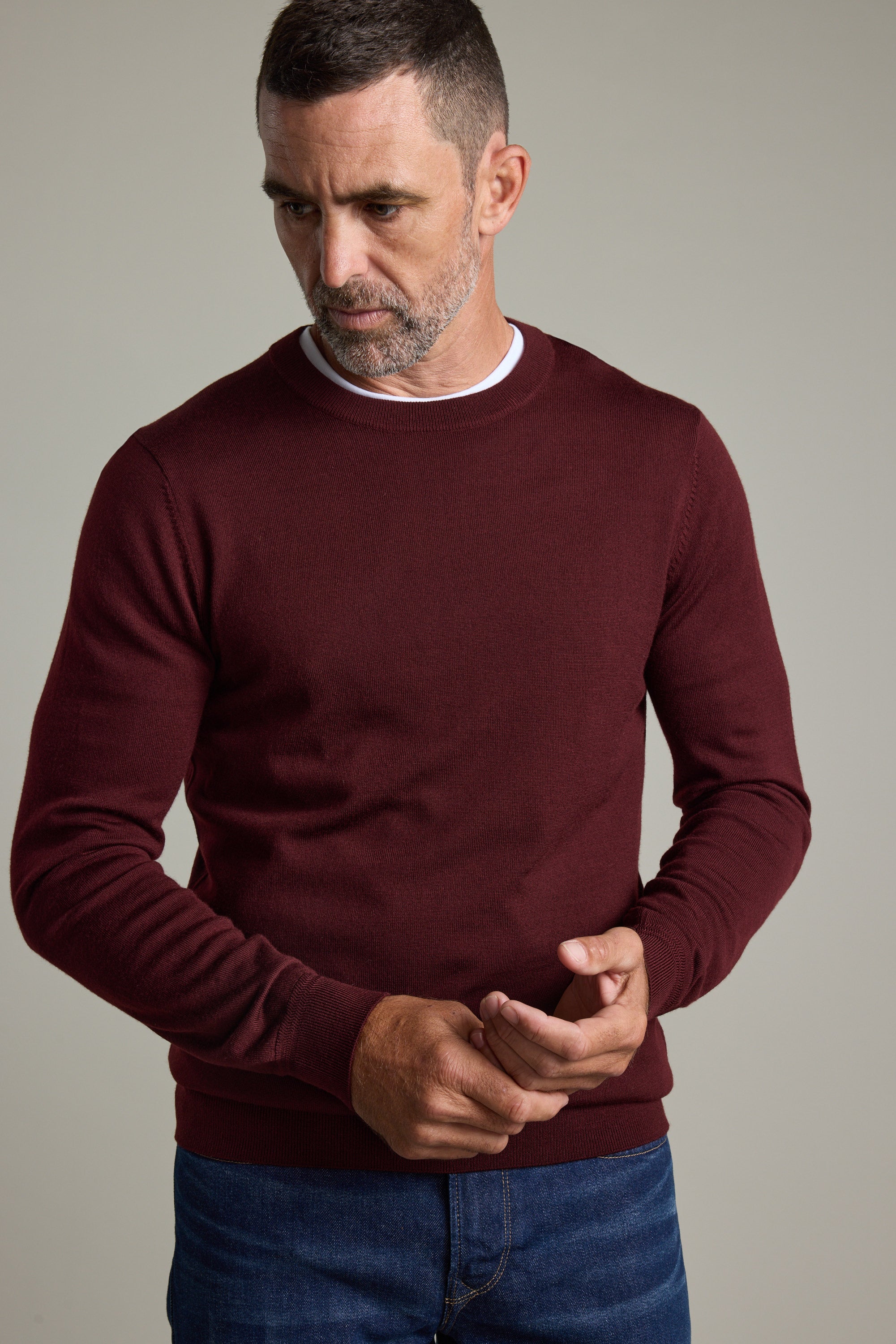 Man in a Barkers Skyland ZQrx Merino Crew and jeans stands against a plain background, looking down.