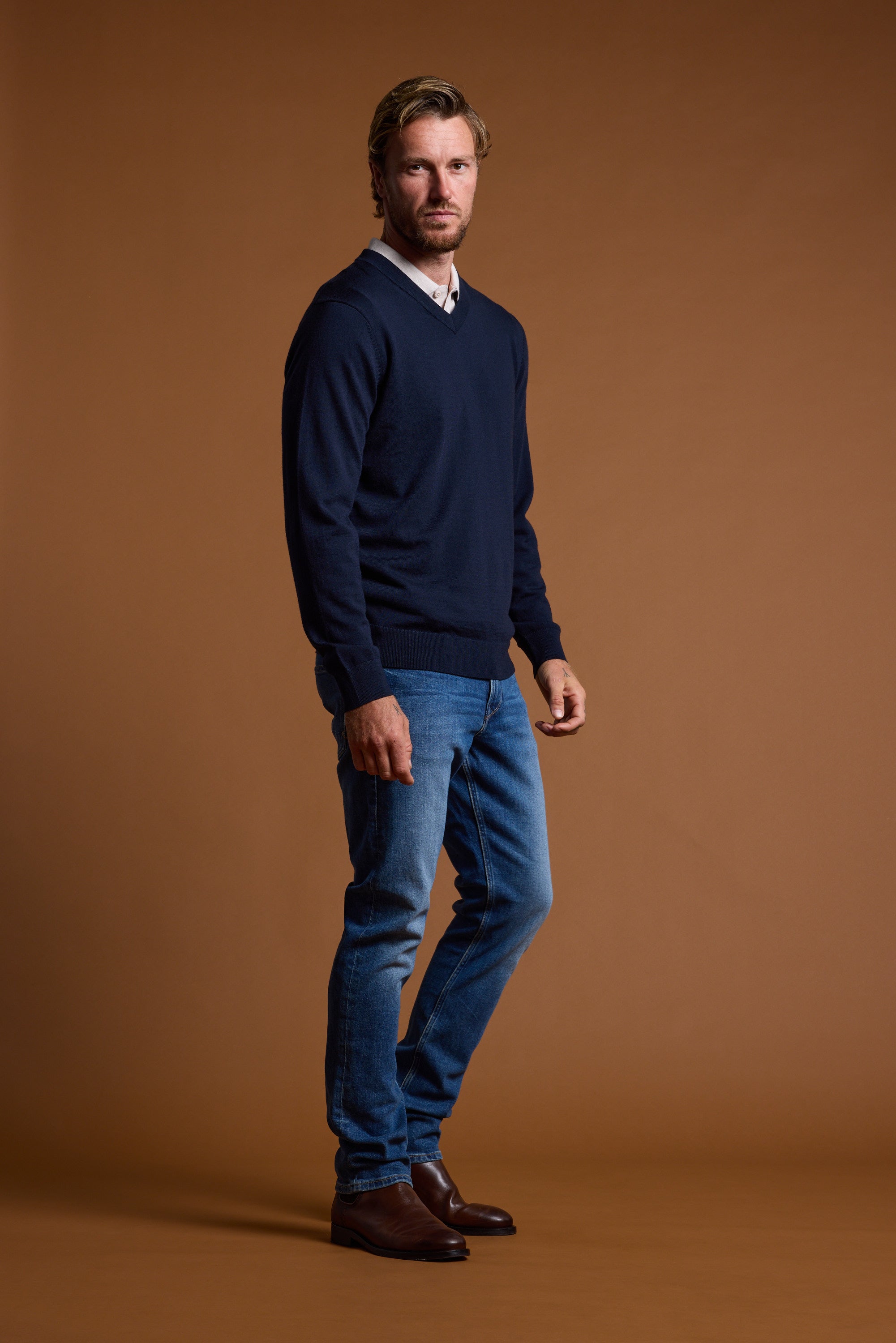 Man wears Barkers Skyland ZQrx Merino Vee sweater, jeans, and boots against a brown background.