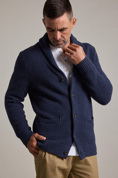 A man with short dark hair and a trimmed beard wears the Barkers Brighton Cotton Shawl Cardi in textured navy blue over a white button-up shirt and beige pants, looking down as he adjusts the cardigan’s collar—an ideal winter layering piece.