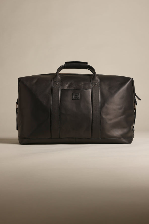 The Barkers Costello Leather Weekender, a black premium leather bag with two handles and a shoulder strap, features a minimalist design and small embossed logo on the front. Displayed against a neutral beige background.
