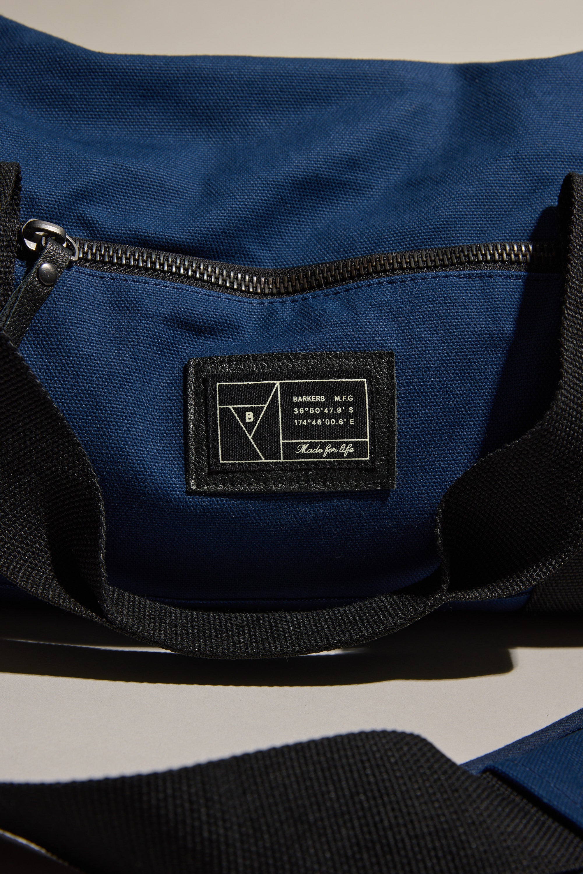 A close-up of the Barkers Canvas Duffle Bag in blue cotton canvas with a black zipper and strap. A stitched black label with white text and geometric logo is seen on the durable outer pocket of this Barkers bag.