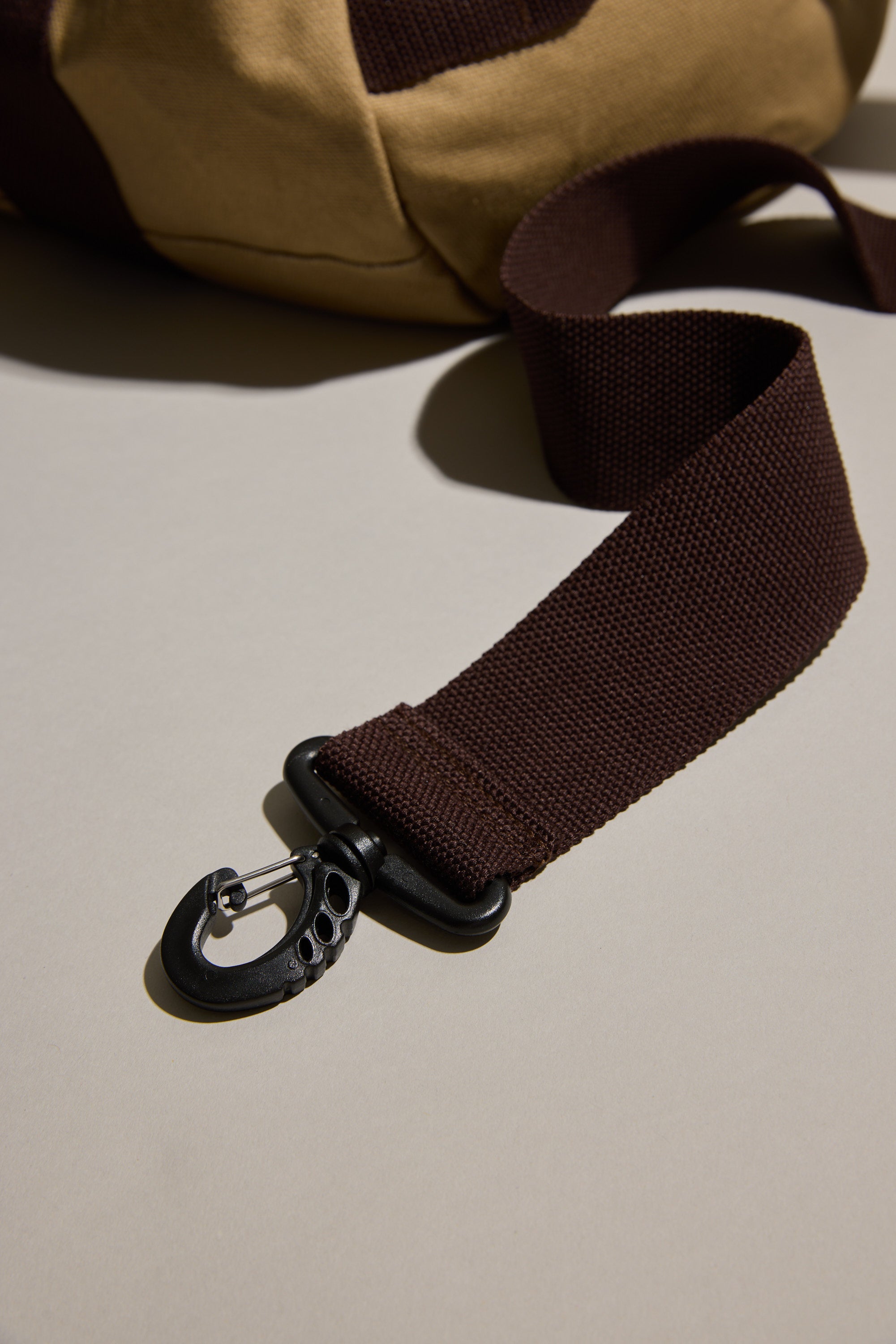A close-up of a dark brown strap with a black carabiner hook, attached to the tan Barkers Canvas Duffle Bag, resting on a light surface—an ideal travel companion from Barkers.