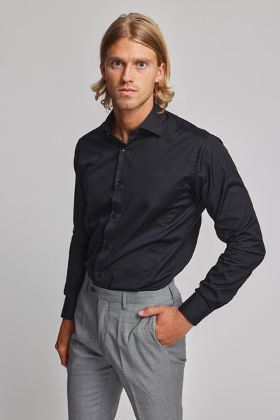 A man with shoulder-length blonde hair models the Barkers Tribeca Reg Cuff Shirt in black, tucked into light gray trousers. He stands against a plain backdrop, one hand in his pocket, gazing neutrally at the camera.