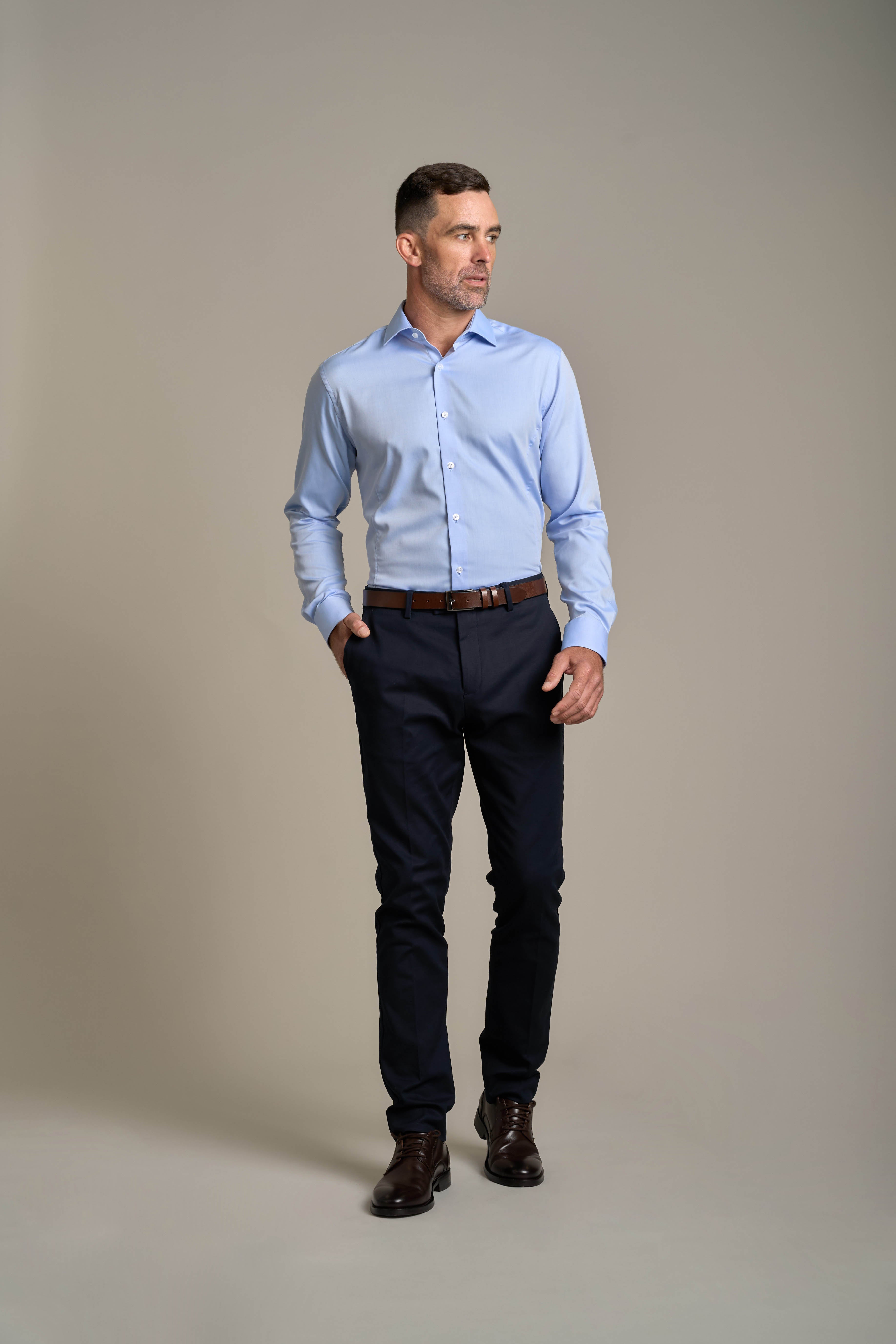 A man in a Barkers Tribeca Reg Cuff Shirt—tailored fit, light blue, wrinkle-free—paired with dark pants and brown shoes stands with one hand in his pocket, looking to the side against a plain light gray background.